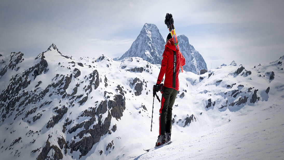 Ein Swiss-Ski-Team-Mitglied mit Slalomstangen auf dem Rücken und einem Einhell Akkubohrschrauber steht auf einem verschneiten Hang mit weitem Blick auf die Alpen.