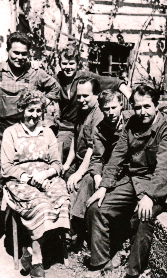 Historic color photo of an elderly woman and five men in work clothes in front of a half-timbered house.