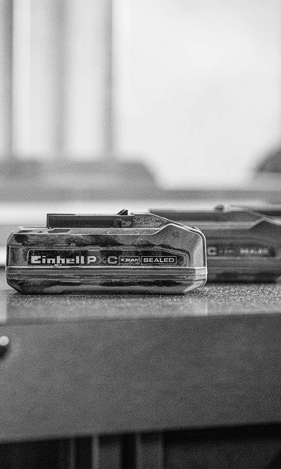 Black and white photo of a dusty Einhell Power X-Change battery on a workbench, label reads: “Einhell PXC 4.0 Ah SEALED”.