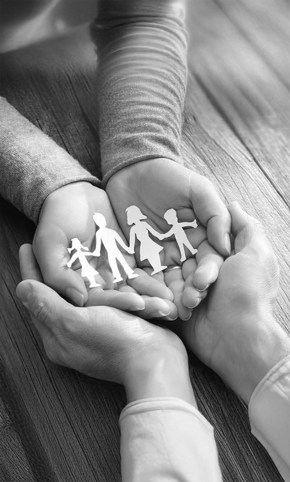 Two pairs of hands hold cut-out paper figures of a family over a wooden table, black and white.