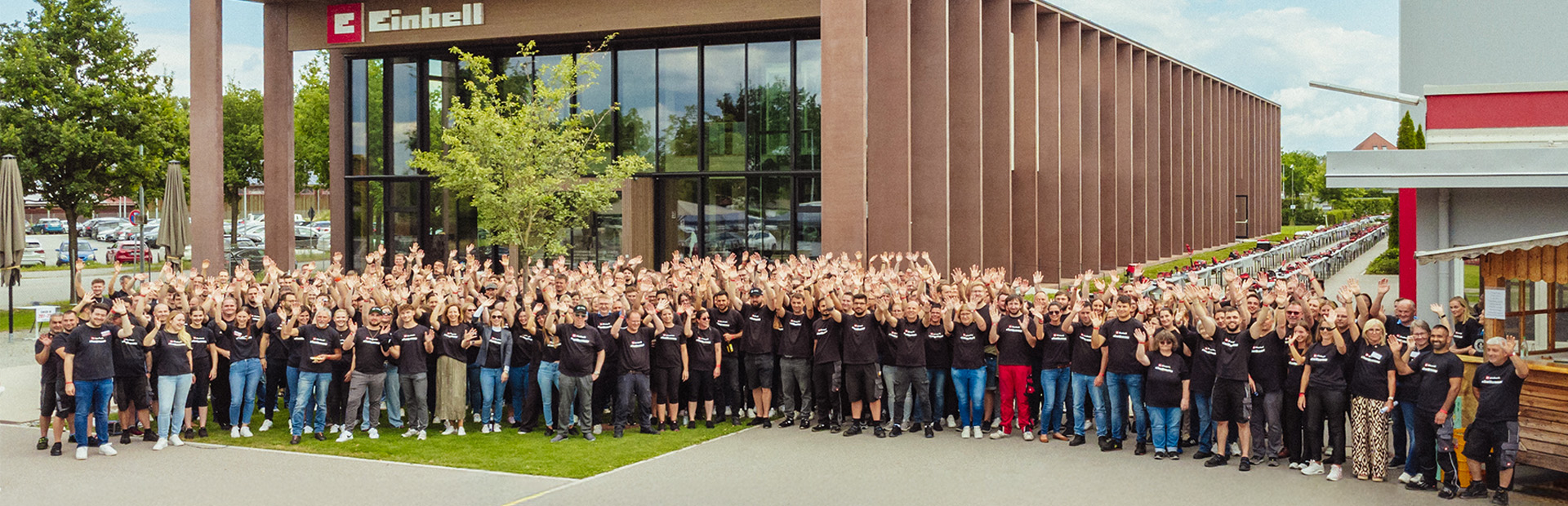 A large group of Einhell employees in black T-shirts waves in front of the modern company building.