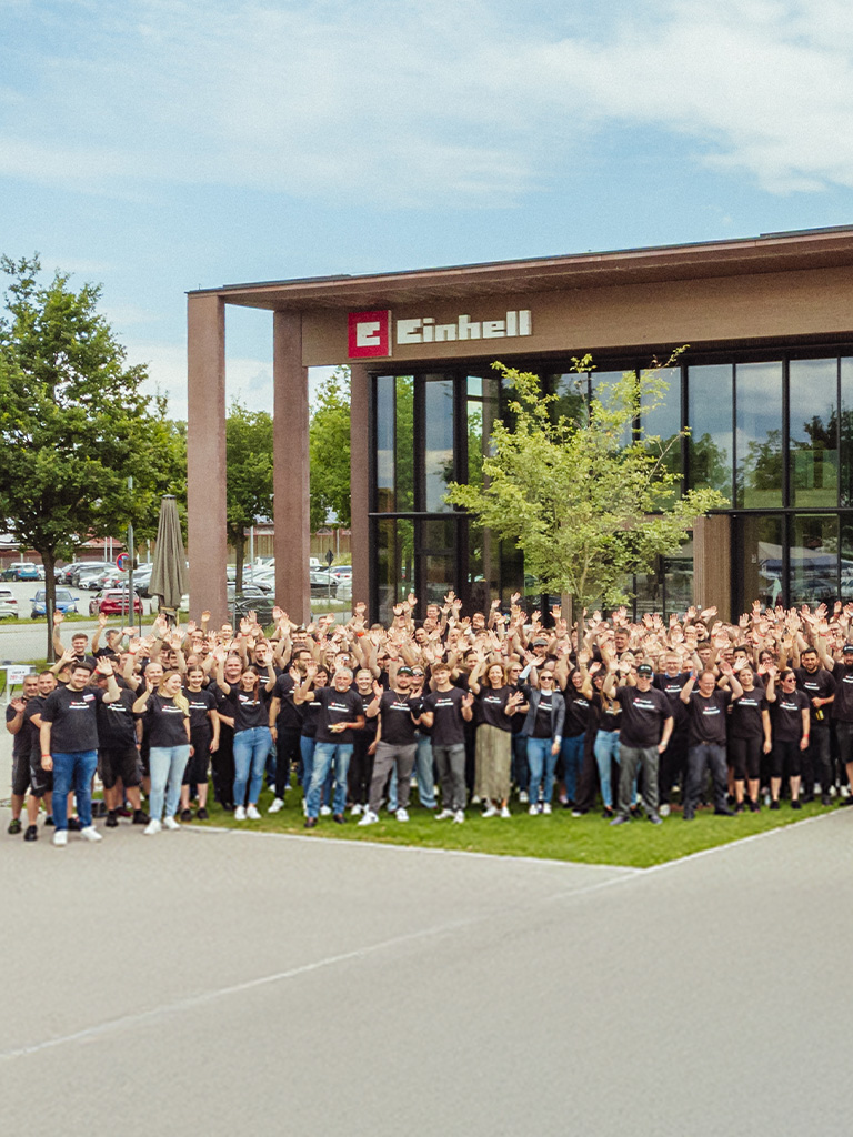 A large group of Einhell employees in black T-shirts waves in front of the modern company building.
