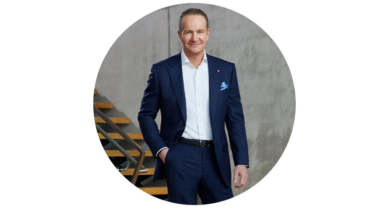 Andreas Kroiss, CEO of Einhell, smiles while standing in a blue suit in front of a concrete wall next to a wooden staircase.