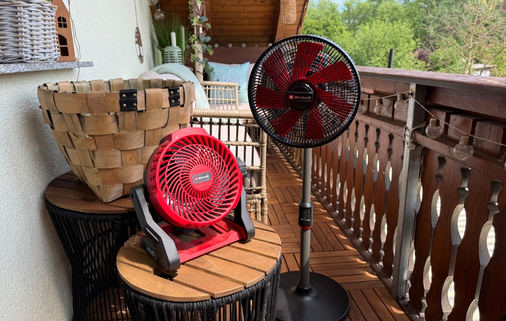 Ein kompakter, roter Einhell Akku-Ventilator steht auf einem kleinen Tisch neben einem größeren Einhell Standventilator auf dem Balkon.
