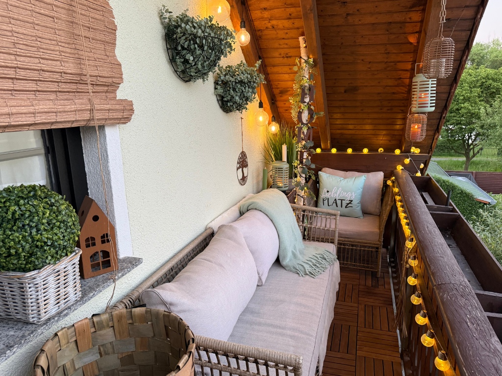 Eine stilvoll eingerichtete Balkon-Lounge am Abend, beleuchtet durch warme Lichterketten, Laternen und Glühbirnen.