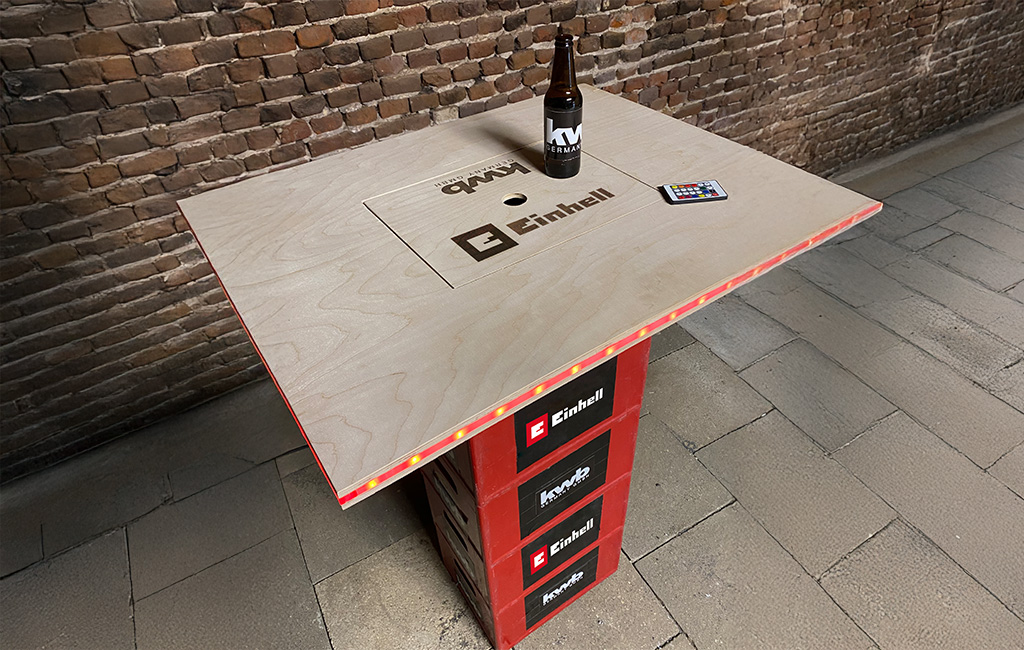 A self-made bar table made of wood with LED strip, placed on a base with Einhell and kwb logos. A beer bottle and remote control are on top. “Einhell” and “kwb GERMANY GMBH” are laser-engraved into the tabletop.