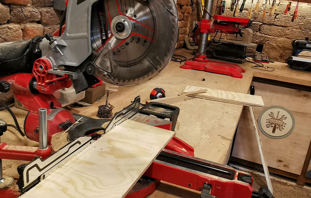 An Einhell miter saw cutting plywood in a woodworking workshop, with red tools on the workbench.