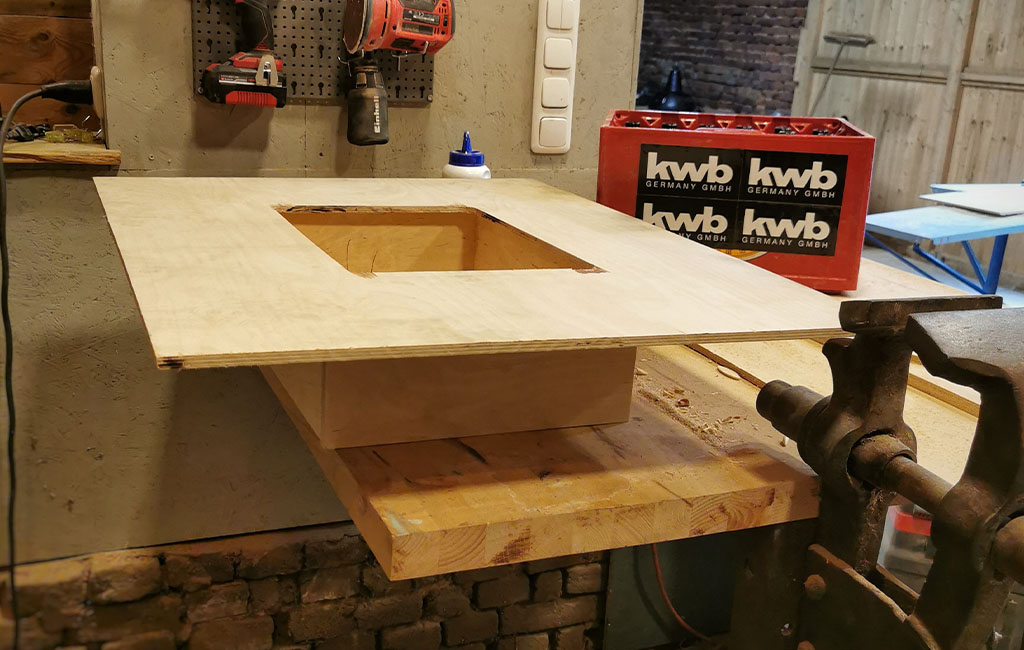 A wooden board with a square cut-out rests on a wooden box. In the background, a tool wall and a kwb drink crate.