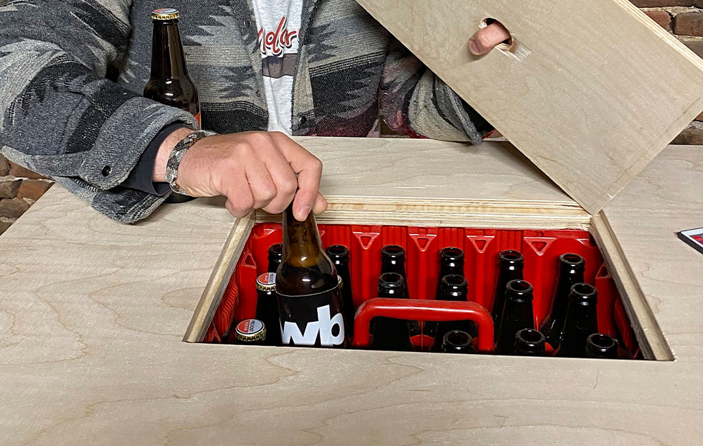 A person lifts the center cover of the table and reaches for a beer bottle inside a red crate.