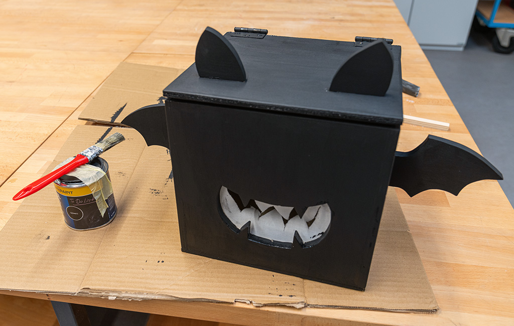 Fertige schwarze Fledermausbox aus Holz mit Ohren, Flügeln und einem weißen Ausschnitt in Form eines gezackten Mauls, daneben eine Farbdose und ein Pinsel.