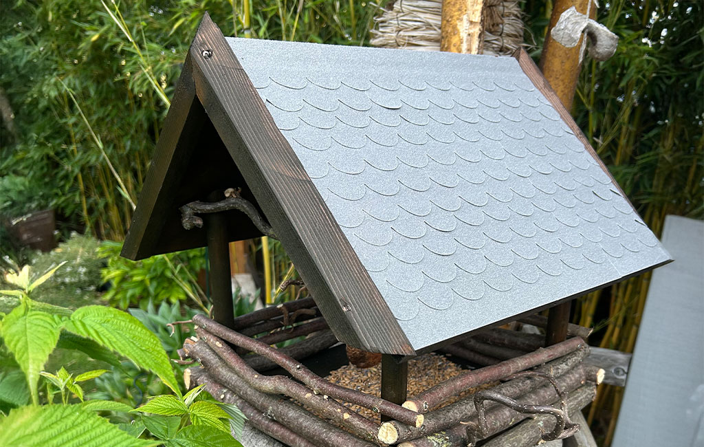Vogelhaus aus Ästen, Holz und grauem Dach steht auf einem Baumstamm im Garten.