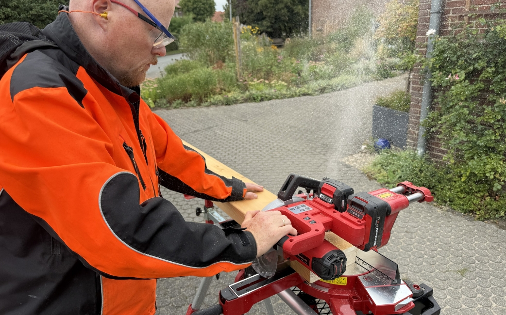 A person in orange and black workwear operates a red power tool on a stand outdoors.
