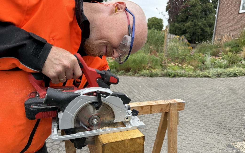 A person wearing protective eyewear uses a handheld circular saw to cut a wooden board supported on a wooden stand outdoors.