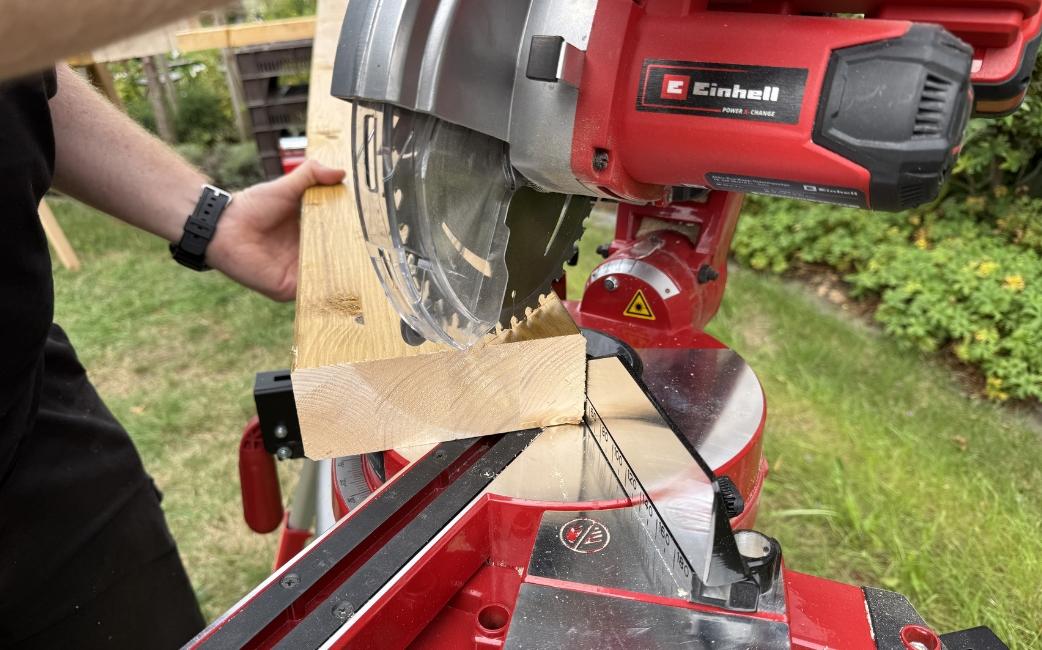 Close-up of an Einhell mitre saw cutting a wooden board, with a hand holding the workpiece in place.