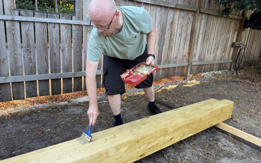 A person applies wood treatment to a wooden beam with a brush in a garden.
