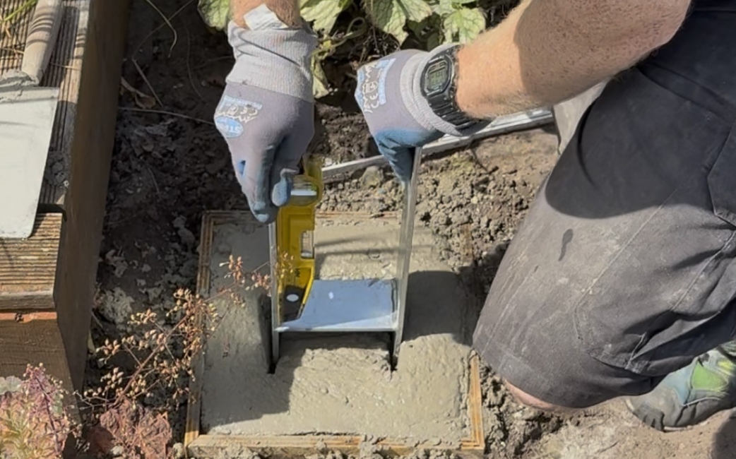 A person wearing gloves uses a yellow hand tool to work on a metal bracket set in fresh concrete beside a wooden structure.