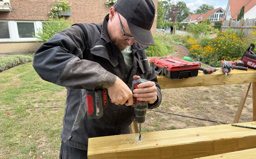 A person in workwear drills a hole in a wooden beam with a cordless drill outdoors.