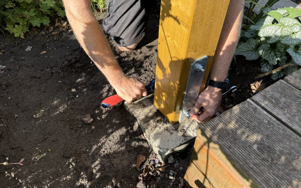 A person fastens a wooden post to a concrete base using a metal bracket and a hand tool.