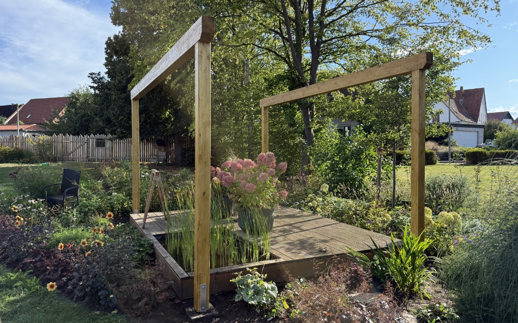 Completed wooden pergola frame on a raised garden platform, surrounded by plants.