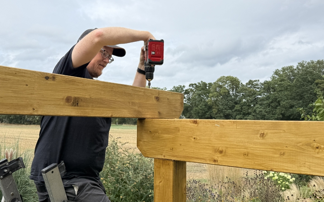  A person uses a cordless drill to fasten a wooden beam to a pergola frame outdoors.