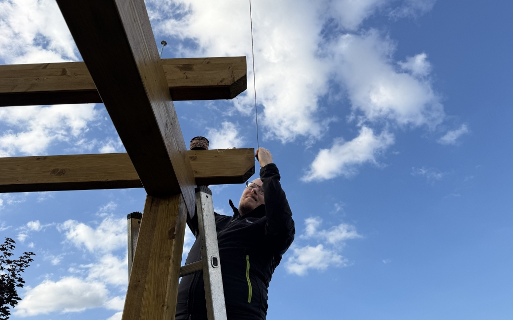 A person on a ladder installs a wooden beam on the upper part of a pergola frame.
