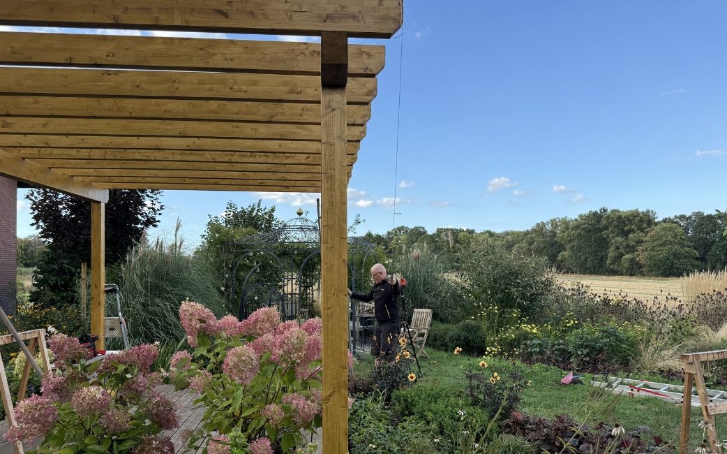 Completed wooden pergola with a slatted roof above a garden seating area, surrounded by flowering plants and greenery.