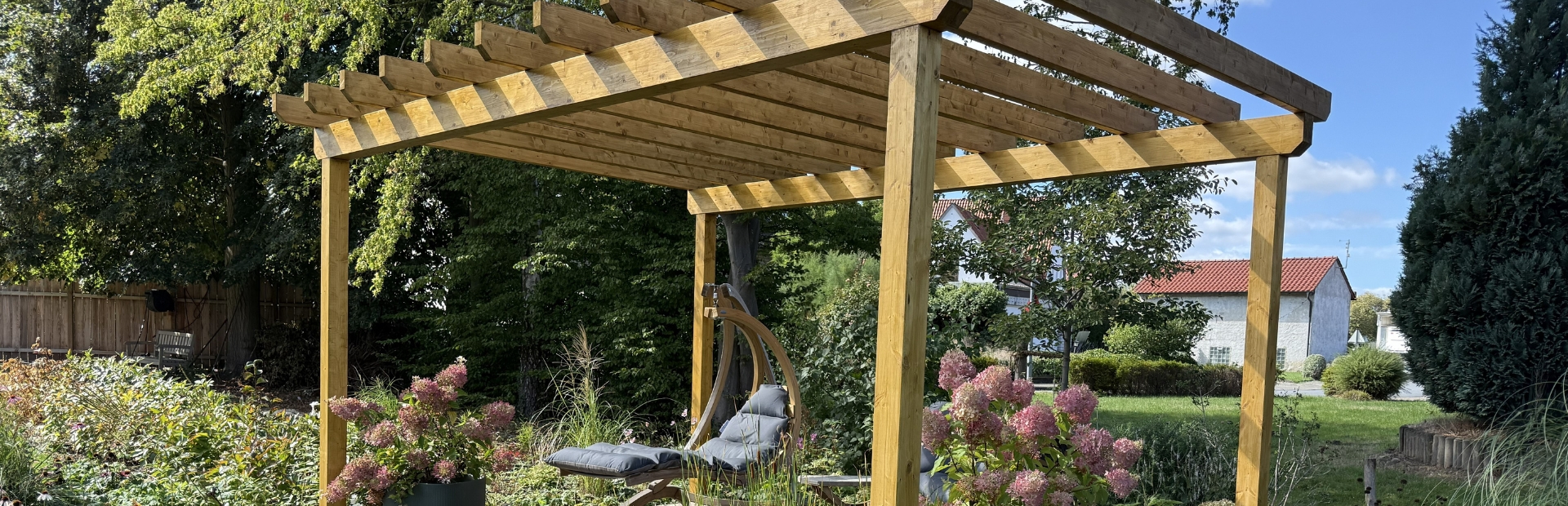 Wooden pergola with a seating area, potted hydrangeas and flowering plants in a garden.