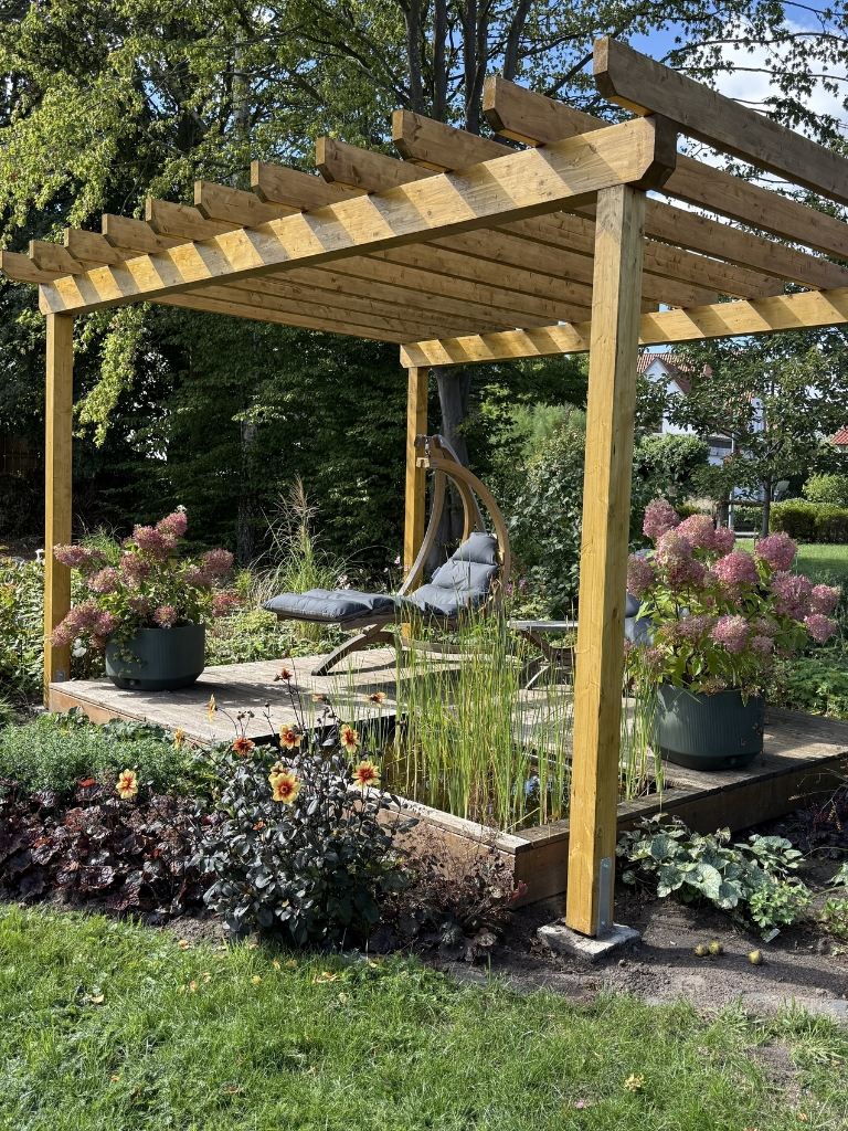 Wooden pergola with a seating area, potted hydrangeas and flowering plants in a garden..