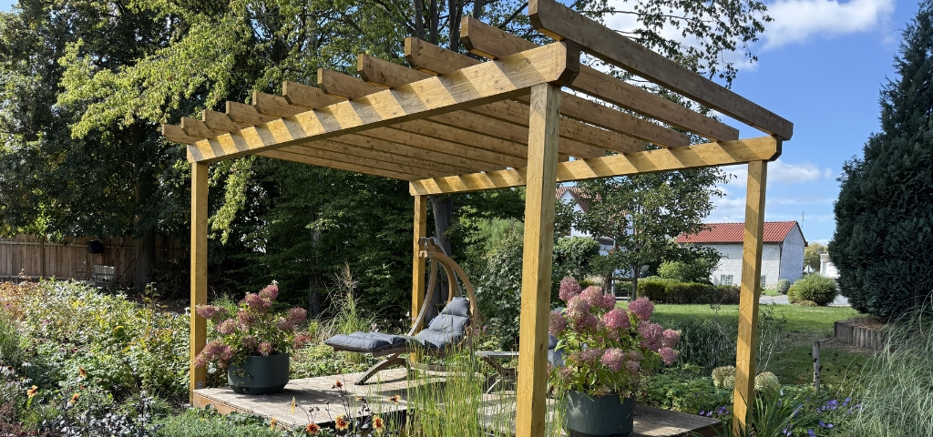 Wooden pergola with a seating area, potted hydrangeas and flowering plants in a garden.