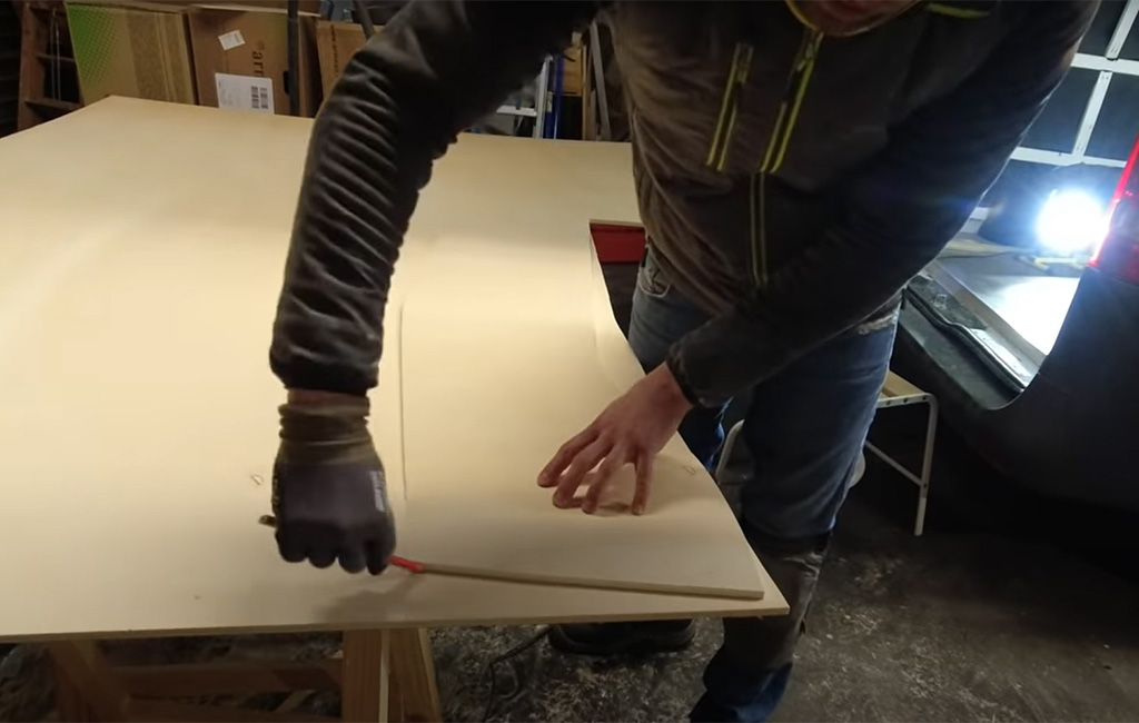Man traces a curved shape onto a plywood sheet using a pencil and a flexible wooden slat.