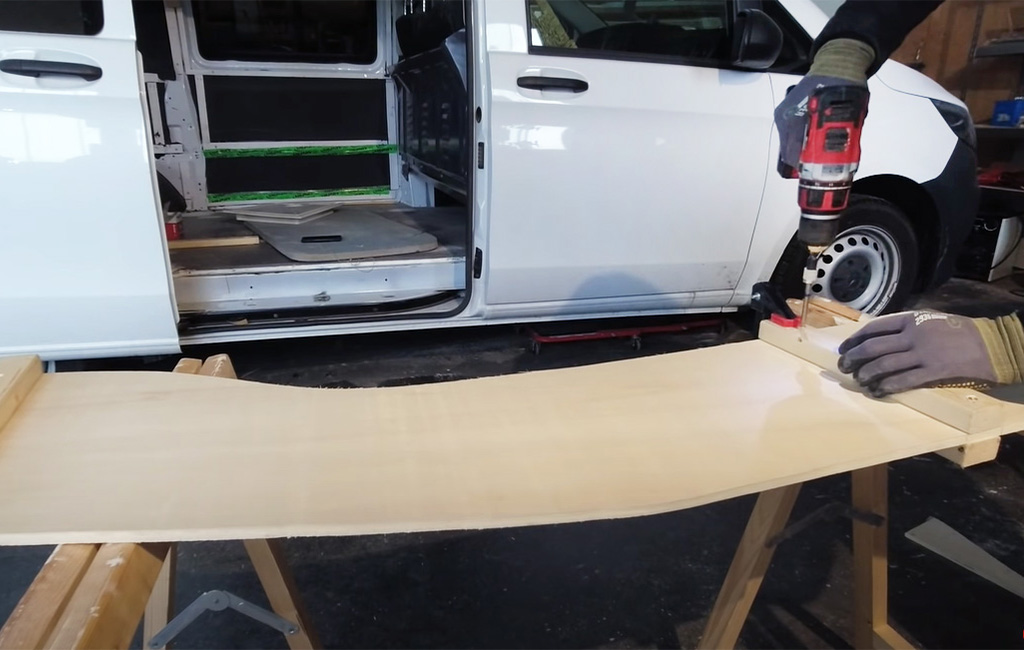 Man drills a curved wooden board with a cordless drill in front of a white van.