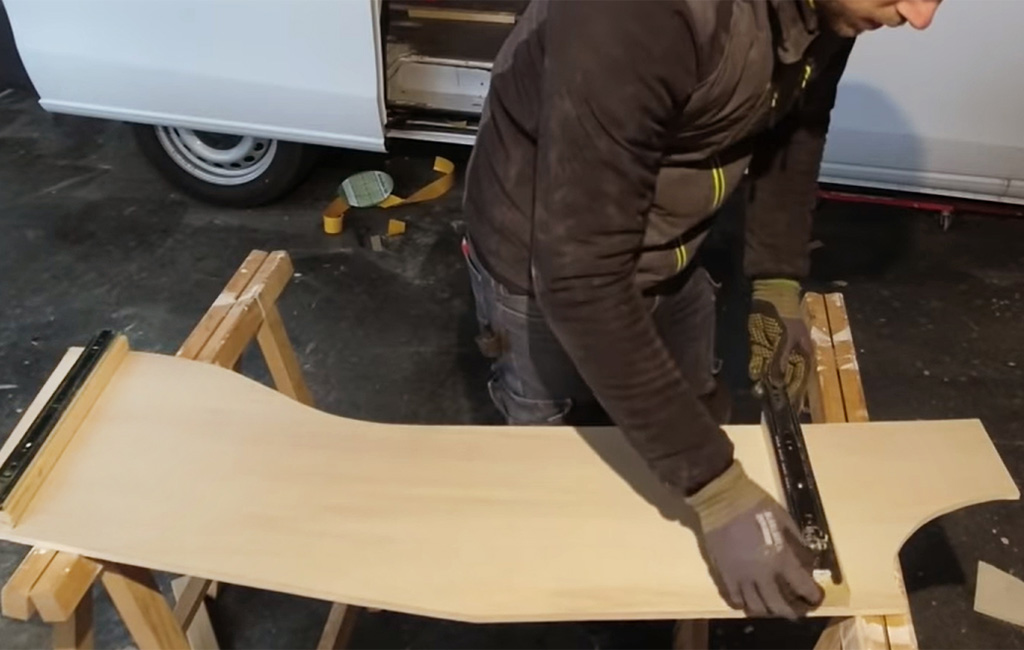 Man mounts a metal drawer slide onto a curved wooden board in front of a camper van.