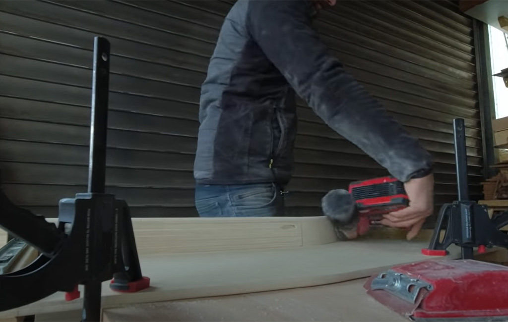 Man sands a wooden board clamped to a workbench using a red cordless random orbital sander.