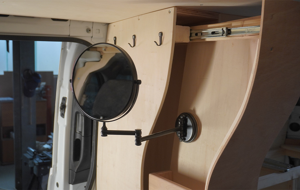 Round mirror with telescopic arm mounted on a wooden panel with hooks inside the camper.