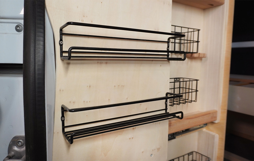 Two black wire baskets mounted on a wooden panel at the door side of the camper cabinet.