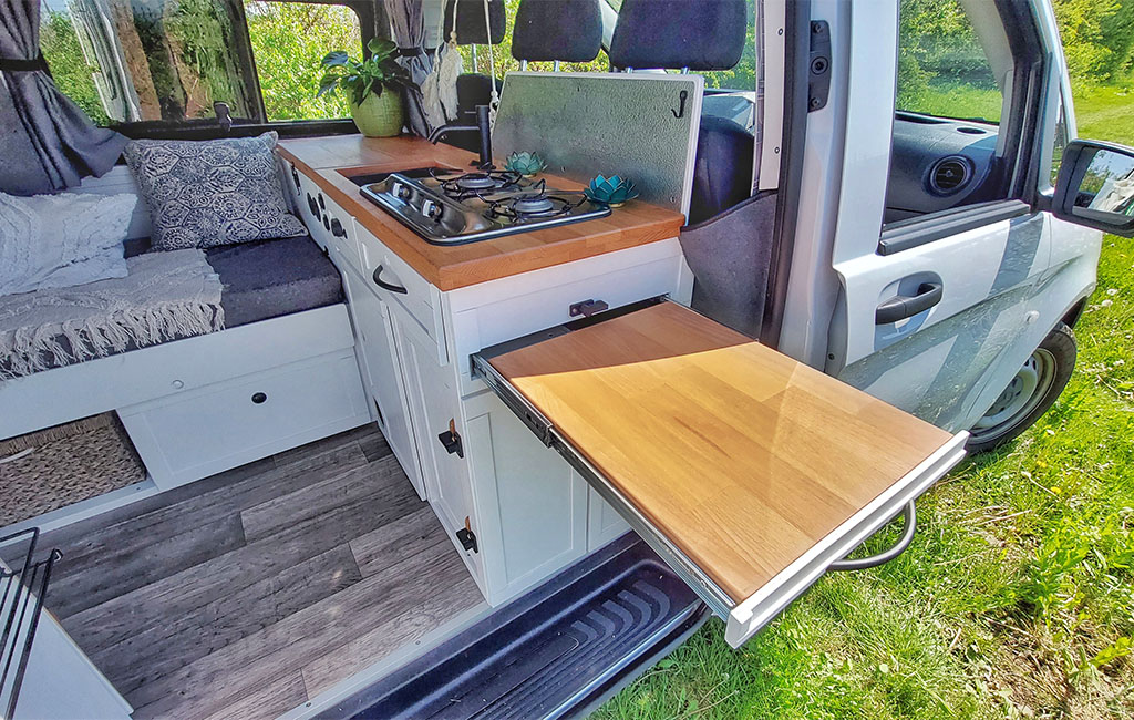 Open camper side door with fold-out wooden table, white kitchenette, gas stove, and plants.
