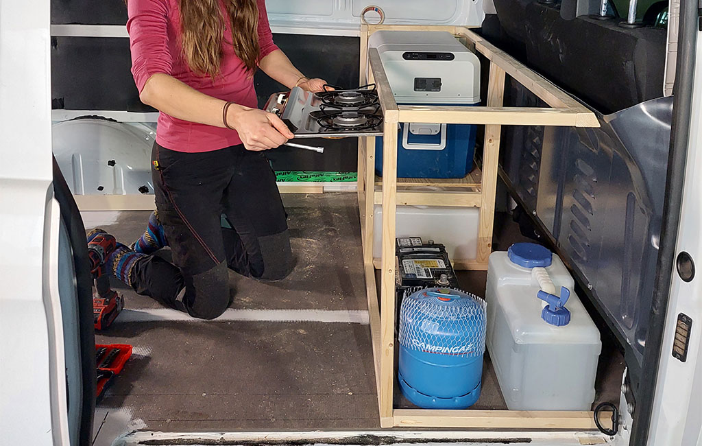 A woman kneels in a van holding a gas stove to be installed in a wooden frame.
