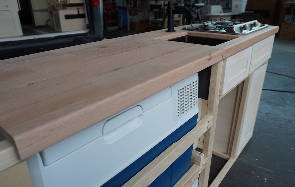 Kitchen unit under construction with wooden countertop, cooler below and open wooden shelving.