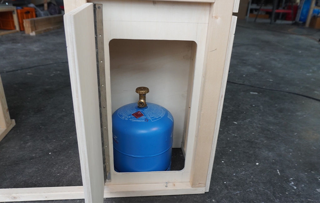 A blue gas bottle is stored in an open wooden compartment of a kitchen unit.