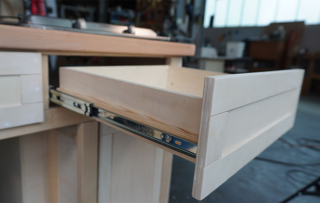 Open drawer made of light wood in a kitchenette with metal sliding rails.