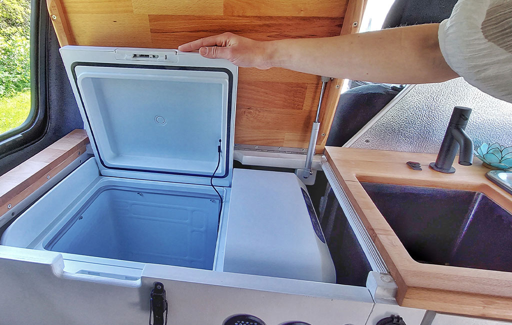 A person lifts the lid of a built-in cooler in a camper kitchen next to a sink.