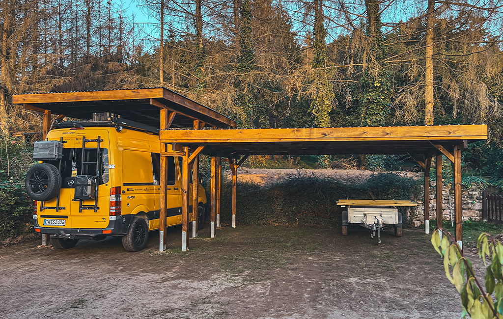 Ein gelber Van und ein Anhänger stehen unter einem großen, offenen Holz-Carport.