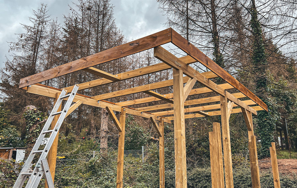 Ein Carport-Rahmen aus Holz mit Streben und Leiter, im Hintergrund Bäume.