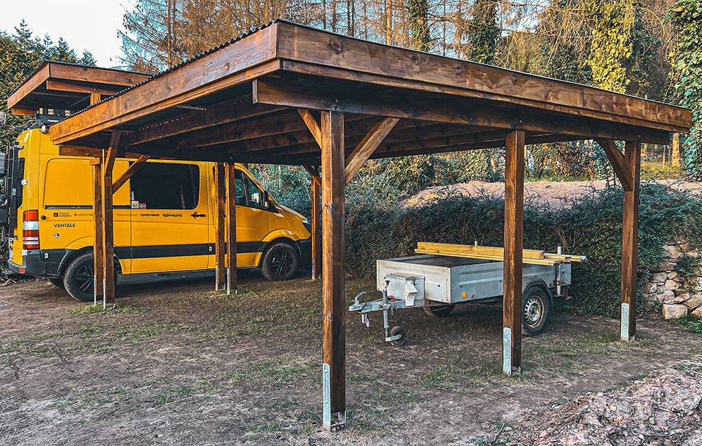 Ein gelber Van und ein Anhänger stehen unter einem fertig gebauten, braun lasierten Holz-Carport.