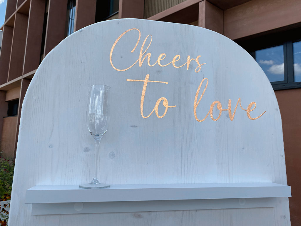 Nahaufnahme des oberen Bereichs einer weiß gestrichenen DIY-Sektwand mit abgerundetem Abschluss, goldfarbener Aufschrift „Cheers to love“ und einem Sektglas auf der Ablage.