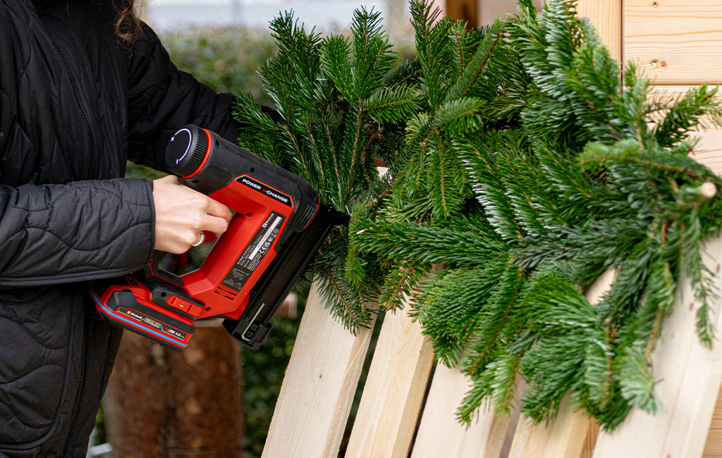 A person attaches fresh fir branches to a wooden pallet using a red Einhell cordless stapler.