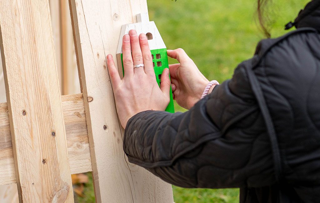 Two hands attach a green and white wooden house to a vertical wooden pallet.