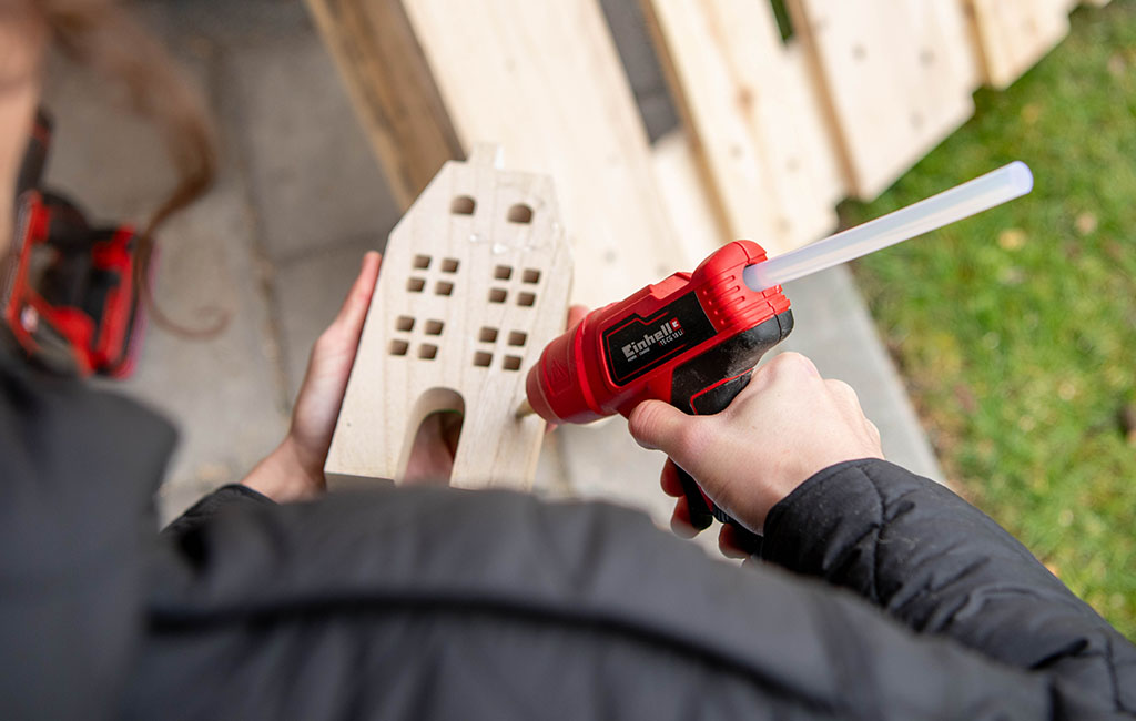 A person applies glue to the back of a wooden house using a red Einhell hot glue gun.