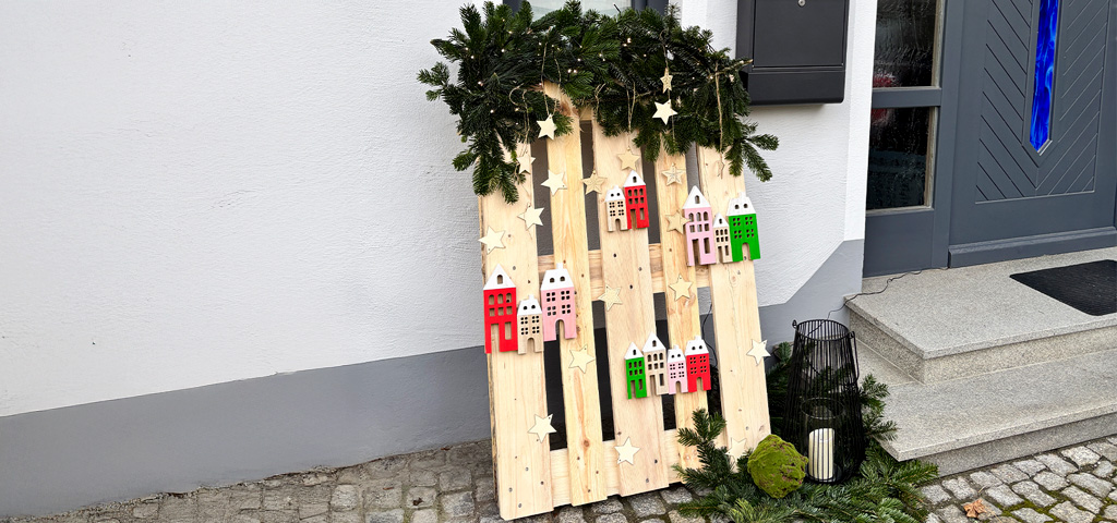 Decorative wooden pallet with stars, wooden houses, and fir branches stands in front of a house entrance on cobblestones.