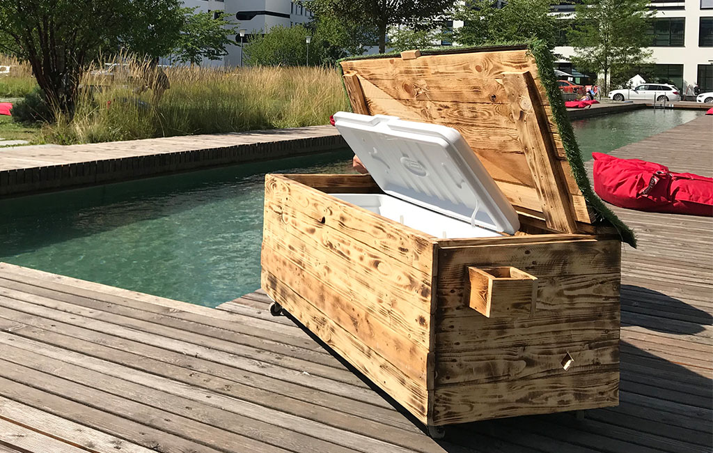 A large wooden chest with an open lid stands on a wooden terrace next to a water basin. Inside is a white cooler.